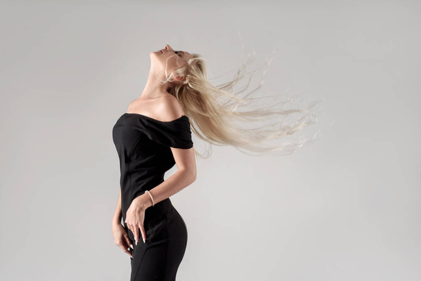 A young slender blonde girl in a stylish black dress shakes her long hair with pleasure. Carefree dancing at a party - 写真・画像