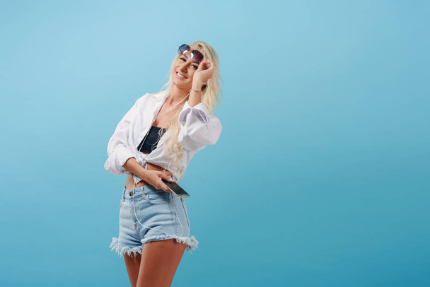 A young slender blonde girl with long hair carefree stands on a blue background and flips music on her headphones. A model in summer clothes and sunglasses smiles and imagines herself on the beach - Foto, imagen