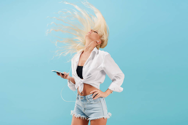 Young slender blond girl with long hair listens to music on headphones and dances, throws back her head. Carefree model in summer clothes having fun on a blue isolated background - Foto, afbeelding