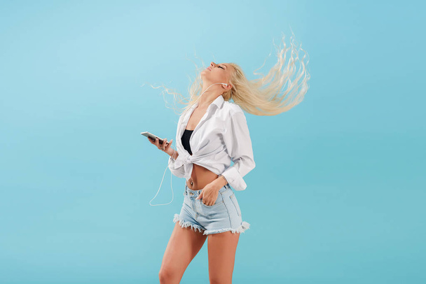 Young slender blond girl with long hair listens to music on headphones and dances, throws back her head. Carefree model in summer clothes having fun on a blue isolated background - Фото, изображение