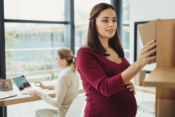 Attentive pregnant woman smiling and taking a book form the shelf - Photo, image