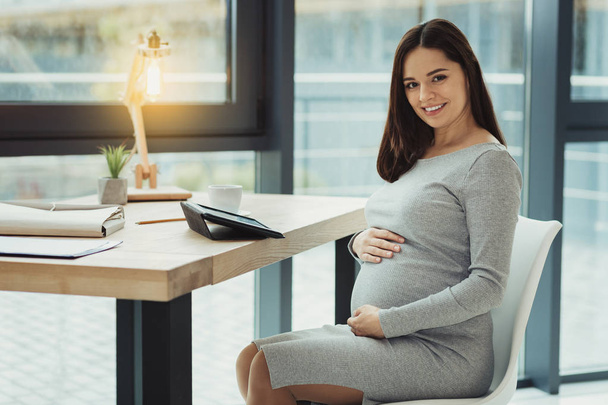 Pregnant young accountant touching her belly and looking happy - Фото, зображення