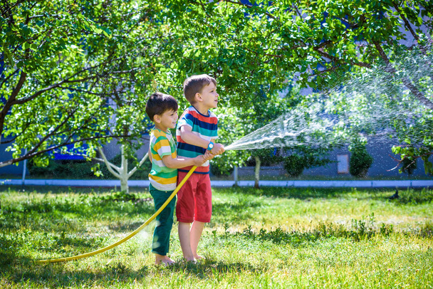 Two little friends playing together and splashing with a garden hose on hot and sunny summer day. Boys having fun with watering fruit garden in village outdoors. Funny holiday leisure water - Photo, Image