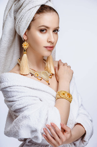 Young woman in a bathrobe and towel on her head, fashion golden jewelry, spa and care portrait, clean natural face, portrait on a background isolated - Фото, зображення