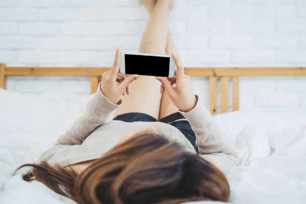 Las mujeres asiáticas felices están utilizando el teléfono inteligente con la pantalla vacía en blanco negro en la cama por la mañana. Mujer asiática en la cama revisando aplicaciones sociales con smartphone. Mujer sonriente navegando por la red con el teléfono celular en casa
. - Foto, Imagen