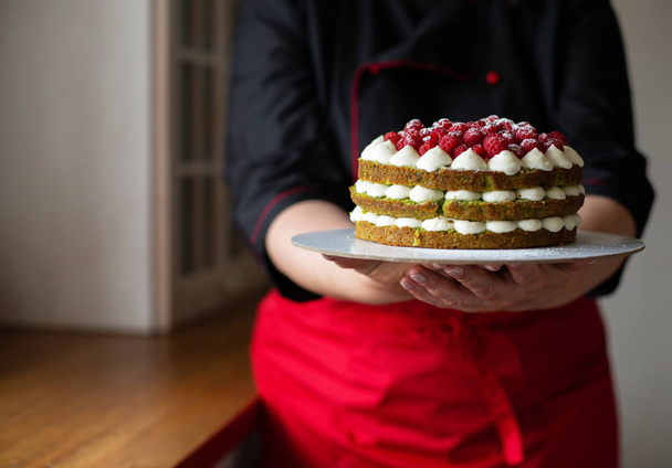 Confeiteiro em uniforme de trabalho vermelho-preto adorna o bolo com bagas na cozinha. Confeiteiro, Bolo, Cozinhar. Espaço de cópia
 - Foto, Imagem
