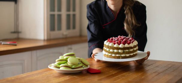 Young Confectioner in  Black  Work Uniform Holding the Cake with Berries  in the Kitchen. Confectioner, Cake, Cooking. - 写真・画像