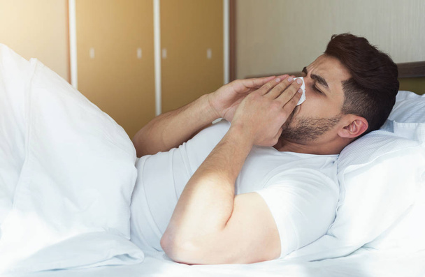 Young man sneezing and covering nose with tissue lying on bed - Photo, Image