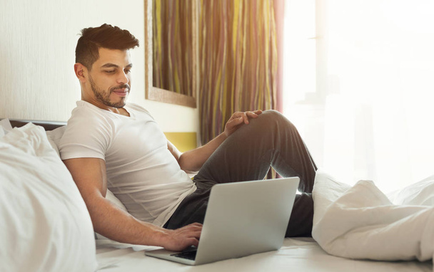 Jeune homme utilisant un ordinateur portable tout en étant assis sur un lit
 - Photo, image