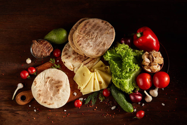 Plain pita on a cutting board with fresh vegetables and cheese on a wooden background, flat lay, selective focus. - Photo, image