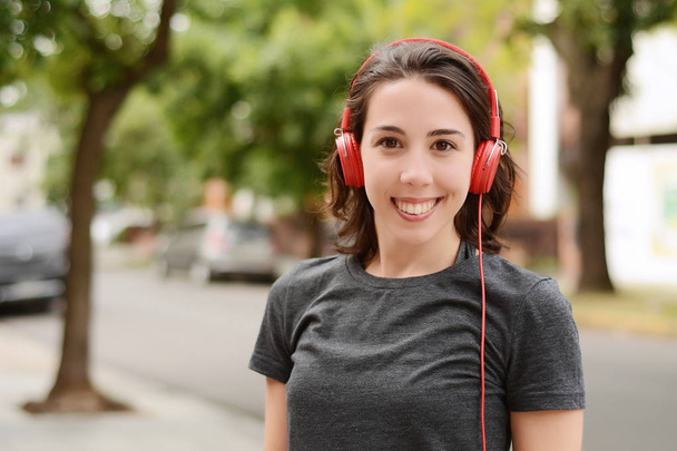 Portrait of young beautiful woman with red headphones listening music. Outdoors. Urban concept. - Фото, изображение