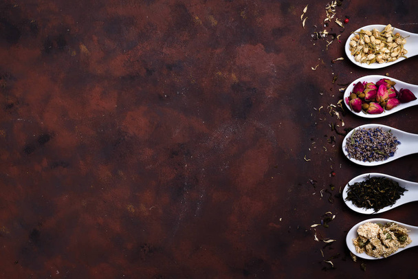 Сушеные цветы жасмина и розы, лаванды, белого и черного чая в глиняных ложках на каменном фоне.
 - Фото, изображение
