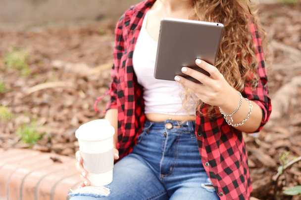 Young student girl with a tablet and a disposable coffee cup sitting on the park in autumn. - 写真・画像