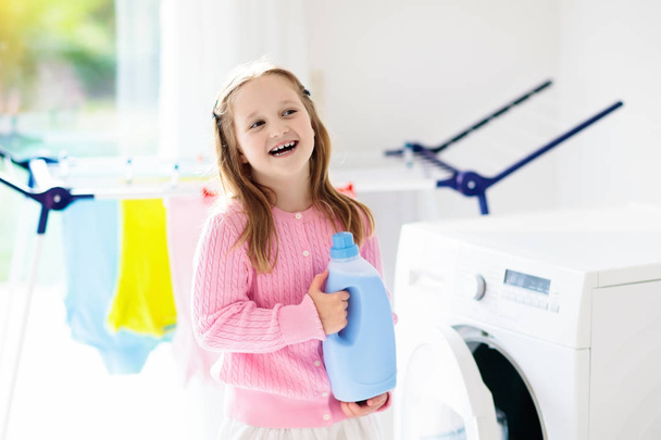 Child in laundry room with washing machine or tumble dryer. Kid helping with family chores. Modern household devices and washing detergent in white sunny home. Clean washed clothes on drying rack.  - Foto, Imagem