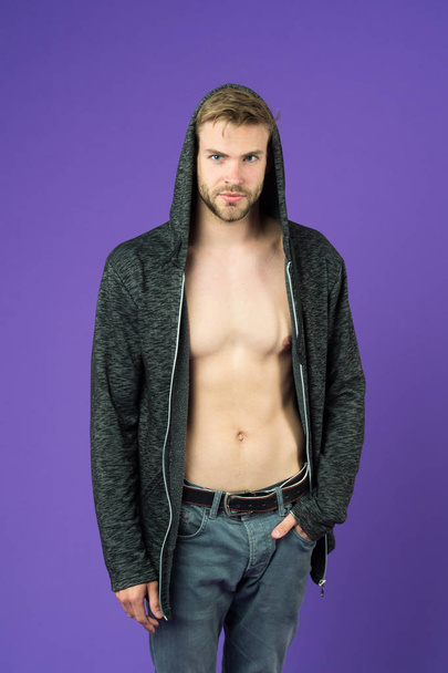 Man with bare torso wearing hoodie. Hipster with trimmed stylish beard in gray jeans. Guy with perfect muscular body. Sexy handsome model isolated on purple background. Street style youth fashion - Valokuva, kuva