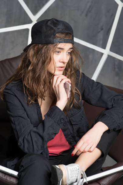Female hipster in oversize black suit. Model with thoughtful look sitting on leather couch. Girl wearing jacket, sneakers and cap backwards. Youth street fashion concept. Woman holding new smartphone - Zdjęcie, obraz