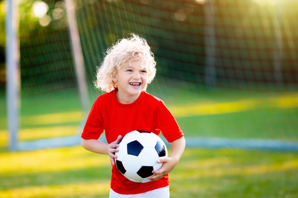 Kids play football on outdoor field. Children score a goal at soccer game. Little boy kicking ball. Running child in team jersey and cleats. School football club. Sports training for young player. - Фото, изображение