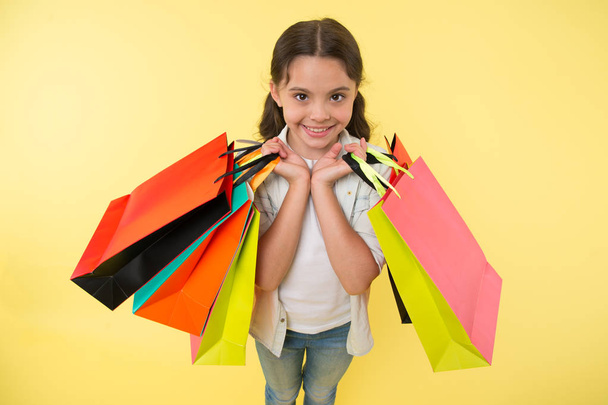 Fashion expert. Child cute shopping expert helps carry packages during shopping. Little shop expert. Girl shopaholic likes shopping. Kid girl happy face carries bunch packages yellow background - Fotografie, Obrázek