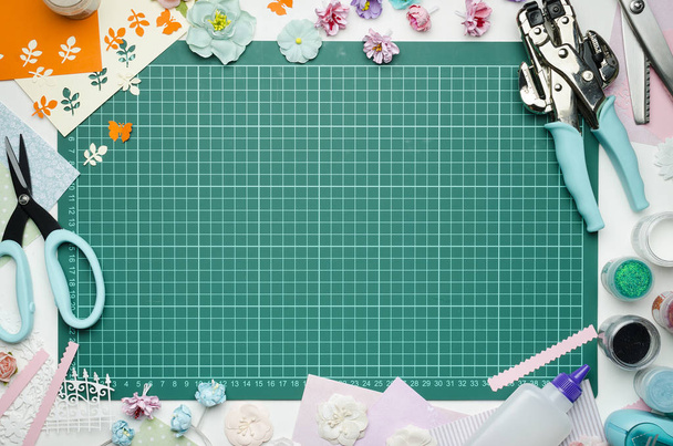 The cutting mat is surrounded by paper flowers, paper, tools and scrapbooking materials. Scrapbooking, top view, empty space in the center - Фото, изображение