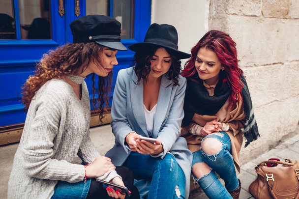 Foto al aire libre de tres mujeres jóvenes mirando el teléfono inteligente en la calle. Chicas hablando y divirtiéndose al aire libre
 - Foto, Imagen