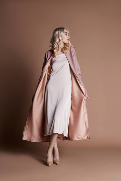 Beautiful blonde woman posing in a pink coat on a beige background. Fashion show clothing, woman with perfect figure, long hair. Trendy fashionable autumn coat on the girl's body - Photo, image