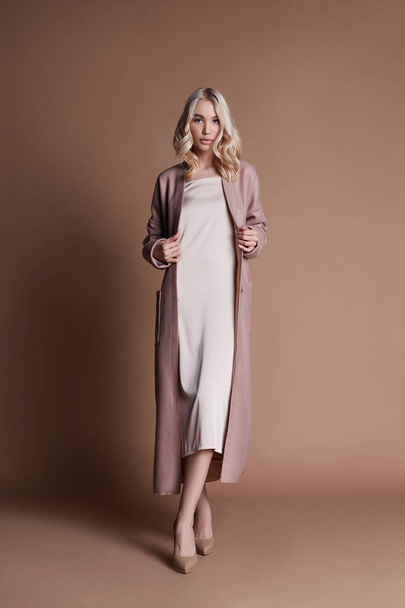 Beautiful blonde woman posing in a pink coat on a beige background. Fashion show clothing, woman with perfect figure, long hair. Trendy fashionable autumn coat on the girl's body - Fotoğraf, Görsel