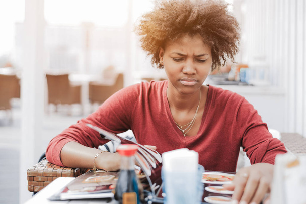 Cute curly African-American girl in street cafe is dissatisfied with an assortment of food in the menu; a young black female is angrily flipping through the menu pages while sitting in a restaurant - Photo, Image
