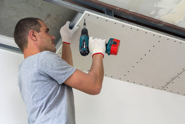 Stavební dělník montuje zavěšený strop se sádrokartonem a upevňuje sádrokarton ke stropnímu kovovému rámu šroubovákem. - Fotografie, Obrázek