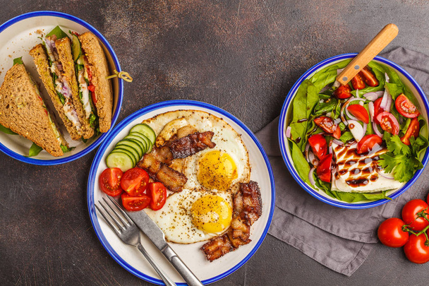 Food flat lay: vegetable salad with goat cheese, sandwiches with cheese and vegetables and fried eggs with bacon. Beautiful bright breakfast on a dark background, copy space - Foto, immagini