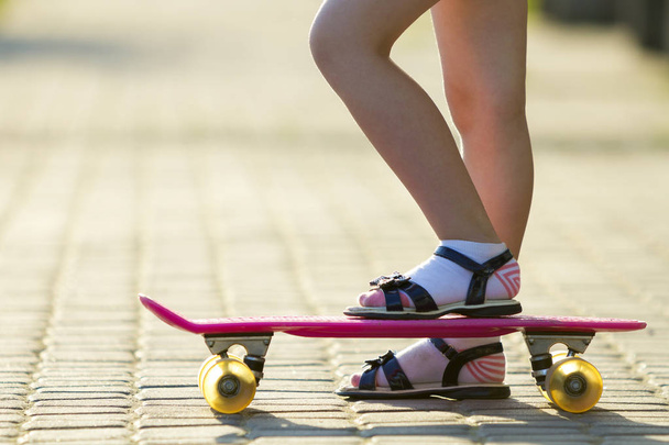 Дитячі тонкі ноги в білих шкарпетках і чорні босоніжки на пластиковому рожевому скейтборді на яскравому сонячному літньому розмитому фоні копіювання простору тротуару. Активність на відкритому повітрі та концепція здорового способу життя
. - Фото, зображення