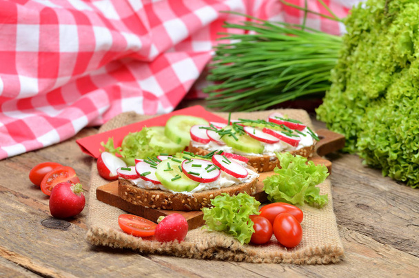 Getreidebrot mit Aufstrich, frischem Rettich, Gurken und Tomaten drauf - Konzept eines gesunden Fitness-Frühstücks oder Snacks, frischer Salat und Schnittlauch im Hintergrund - Foto, Bild