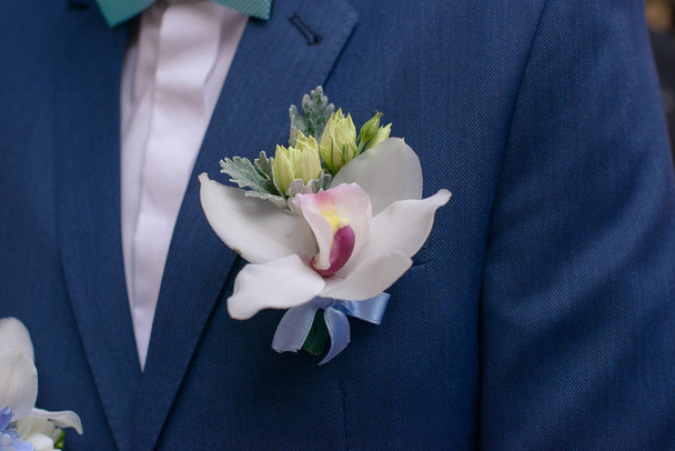 Nahaufnahme von frischer Orchidee boutonniere, die an einen Bräutigam oder eine männliche Gastjacke geheftet ist, ein traditionelles männliches Accessoire für Hochzeiten, das häufig zum Brautstrauß passt oder mit ihm kombiniert wird. - Foto, Bild
