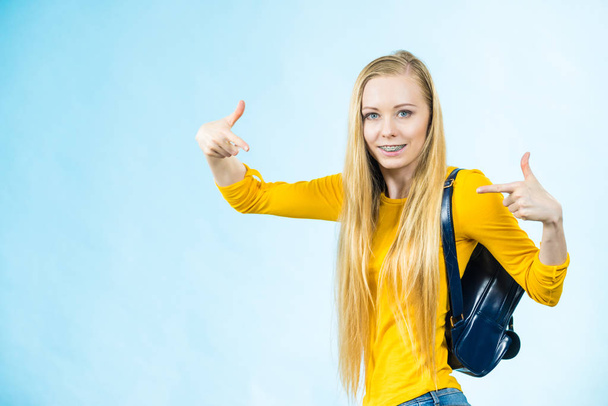 Happy young blonde teenage girl going to school or college wearing backpack pointing at herfelf being proud. Studio shot on blue. - 写真・画像