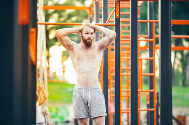 Subject sport street workout Handsome young caucasian man with a bare-chested muscular with long red hair and beard posing outdoor sports ground, gym outdoors. Sexy guy touches his hair - Photo, Image