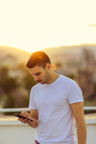 Έφηβο που στέκεται στο μπαλκόνι στο ηλιοβασίλεμα και γραπτών μηνυμάτων στο κινητό του τηλέφωνο - Φωτογραφία, εικόνα