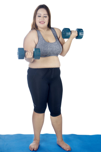 Portrait of Caucasian fat woman lifting two dumbbells while standing on the mat, isolated on white background - Fotografie, Obrázek