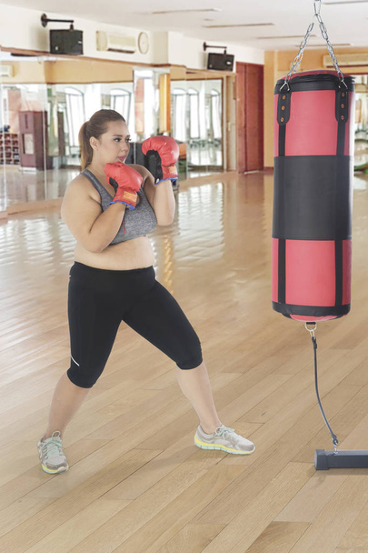 Portrait of a young fat woman exercising with a boxing bag while standing in the fitness center - Photo, Image