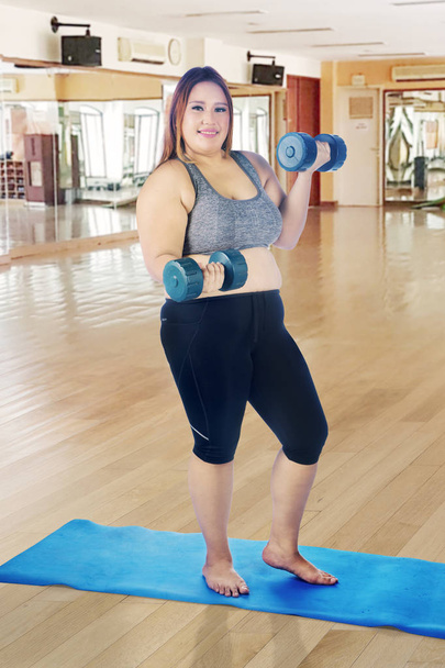 Portrait of happy fat woman exercising with two dumbbells while standing on the mat. Shot in the fitness center - Photo, Image