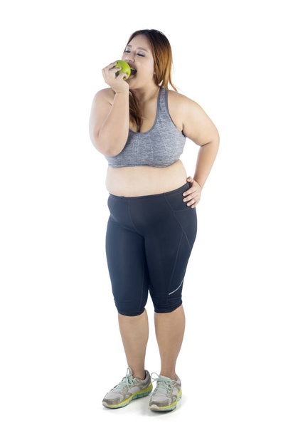 Full length of obese woman wearing sportswear while eating a green apple, isolated on white background - Photo, Image