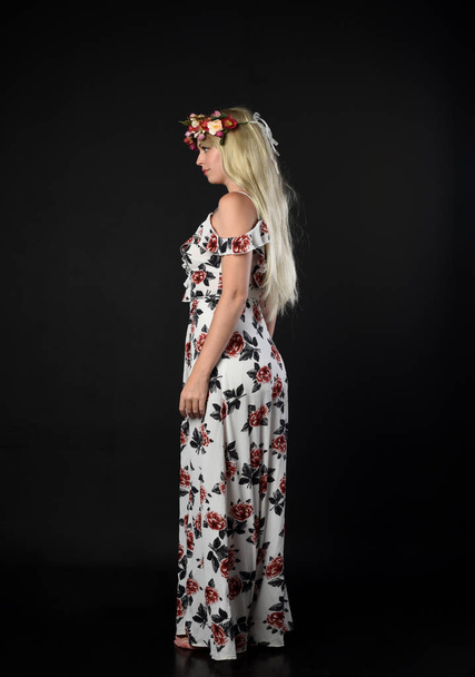 Ολόσωμο πορτρέτο του ξανθιά κοπέλα φορώντας floral φόρεμα, όρθια στάση σχετικά με φόντο μαύρο στούντιο. - Φωτογραφία, εικόνα