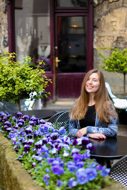 Mujer joven sentada en la cafetería de la calle cerca de flores azules y plantas verdes
. - Foto, Imagen