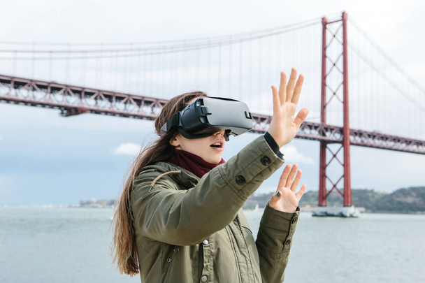 Menina bonita nova vestindo óculos de realidade virtual. Ponte 25 de Abril, em Lisboa, ao fundo. O conceito de tecnologias modernas e seu uso na vida cotidiana
 - Foto, Imagem