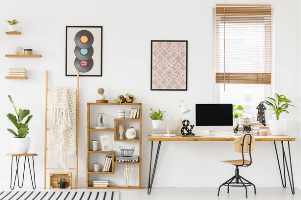 Real photo of a workplace interior with a computer on a desk next to a chair and a shelf with ornaments, posters on a wall and a window with blinds - 写真・画像