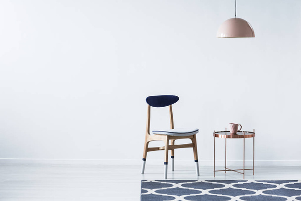 Wooden chair next to copper table under pink lamp in minimal interior with copy space and blue carpet. Place for your poster - Φωτογραφία, εικόνα