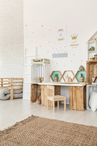 Small wooden chair standing by the desk and crate shelves in white Scandi baby girl room interior with carpet and posters on the wall - 写真・画像