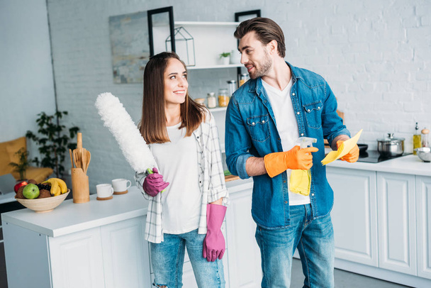 happy couple holding cleaning tools and looking at each other in kitchen - Photo, Image