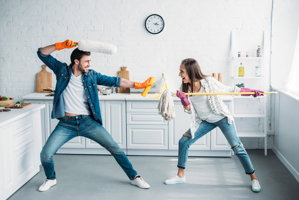 couple having fun and pretending fight with cleaning tools in kitchen - Photo, Image