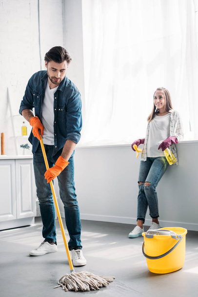 boyfriend cleaning floor in kitchen with mop and girlfriend leaning on kitchen counter - Photo, Image