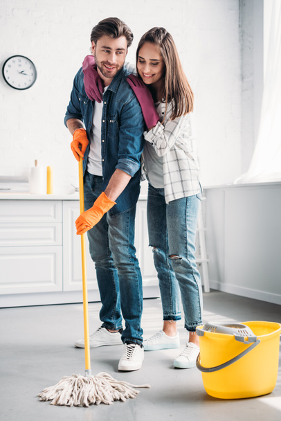 novio limpieza piso en cocina con fregona y novia abrazándolo
 - Foto, imagen
