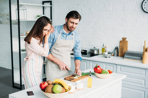 boyfriend cutting onion and crying girlfriend leaning on him in kitchen - Фото, изображение
